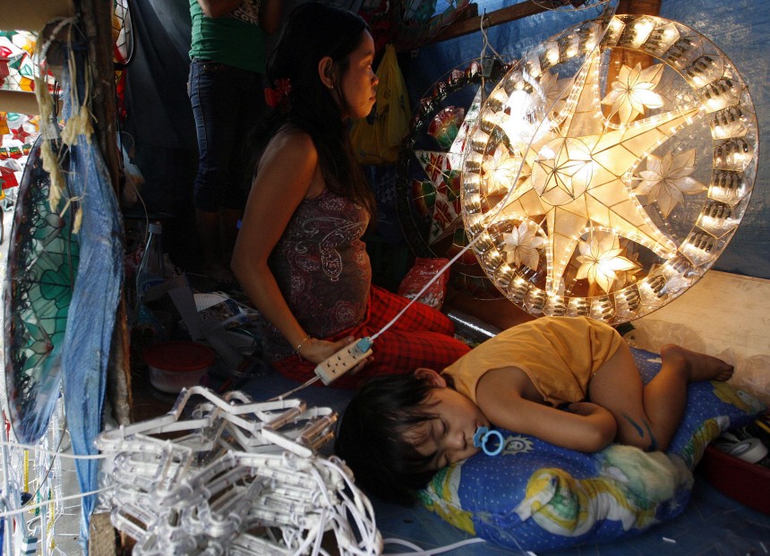 A woman tests the lights of Christmas decorations beside her son sleeping inside their makeshift stall along a busy street in Manila
