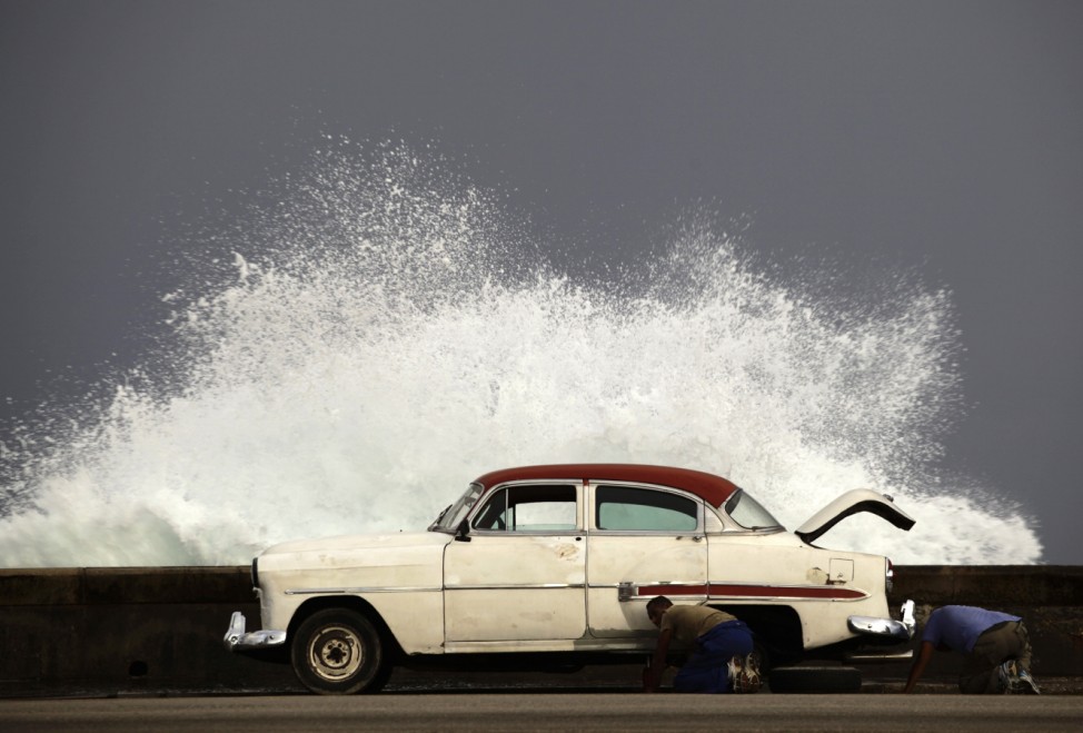 Men change a flat tyre of their car on Havana's seafront boulevard as a wave crashes