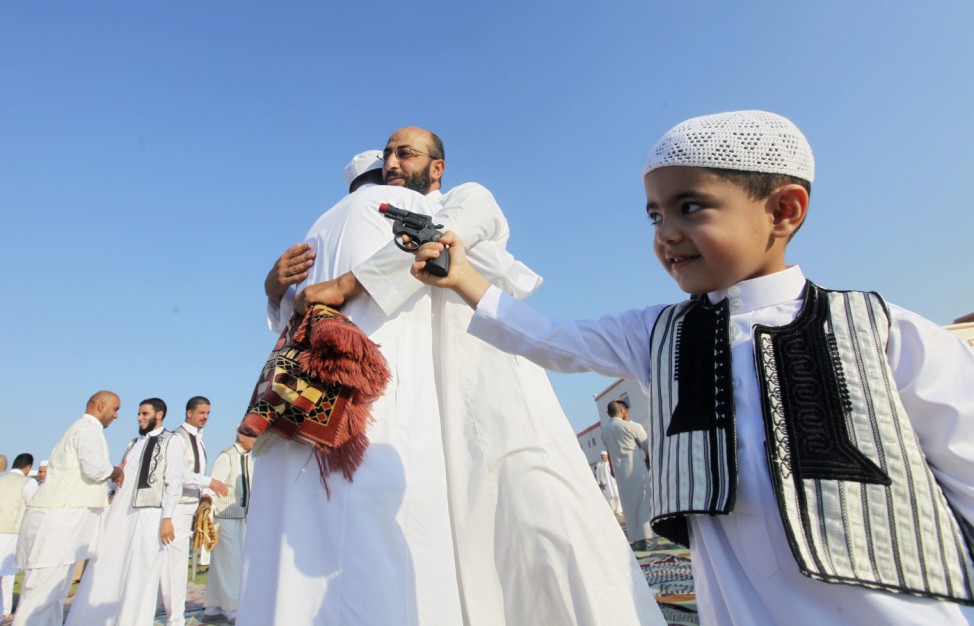 A boy plays with his toy gun near two men hugging after Eid al-Fitr prayers in Benghazi