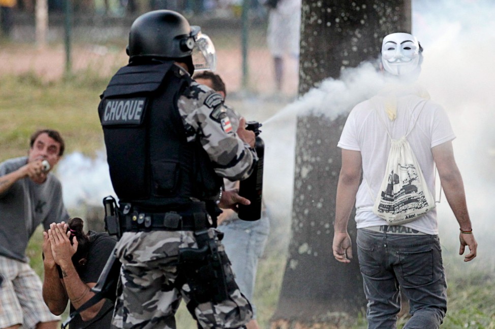 A police officer fires pepper spray at a protester wearing a Guy Fawkes mask on the back of his head to stop a demonstration in Salvador
