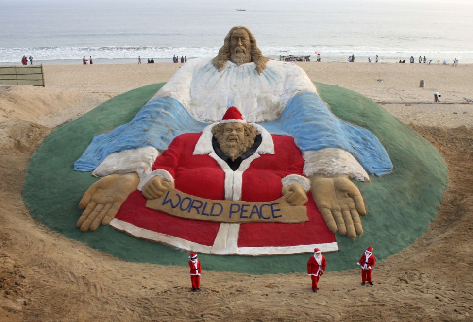 Children dressed in Santa Claus costumes pose in front of a sand sculpture featuring Jesus Christ and Santa Claus created by Indian artist Pattnaik on a beach in Puri