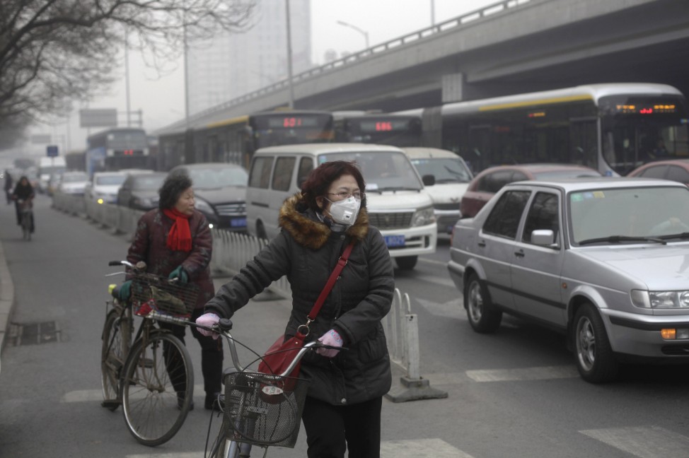 A woman wearing a mask stands besides her bicycle as vehicles stop at a traffic junction on a busy street amid thick haze in Beijing