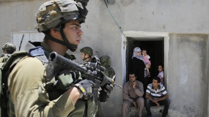 Palestinians sit outside their house as Israeli soldiers patrol near Hebron