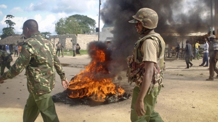 Nach Massaker: Bewaffnete Sicherheitskräfte gehen an einer brennenden Barrikade vorbei, mit der die Dorfbewohner von Kibaoni bei Mpeketoni gegen das Massaker und die mangelnde Unterstützung durch die Regierung protestieren.