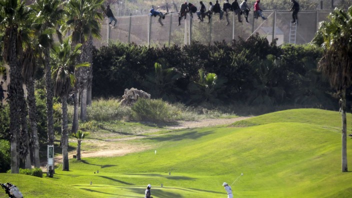 A golfer hits a tee shot as African migrants sit atop a border fence during an attempt to cross into Spanish territories between Morocco and Spain's north African enclave of Melilla