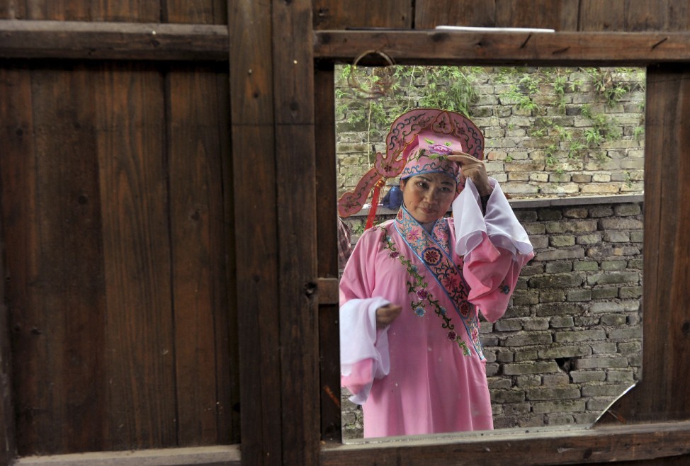 A Chinese folk opera performer in traditional costume adjusts her hat as she is reflected in a mirror placed on a wooden wall, before a performance at a tourism resort in Longyou county