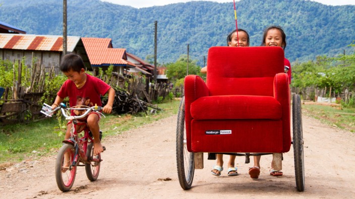 Mit einem Sessel auf Weltreise: Wo er auch hinkommt, er sorgt für Aufsehen: La Silla, der rote Sessel. Hier freuen sich Kinder in Laos über den Besuch.