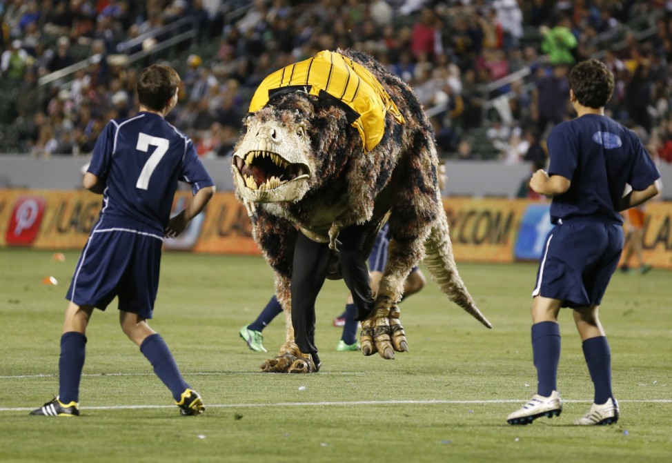 A person in a Tyrannosaurus Rex dinosaur costume runs on the field as young boys play an exhibition soccer match during halftime of the MLS soccer game between the Los Angeles Galaxy and Seattle Sounders FC in Carson; Dinosaurs