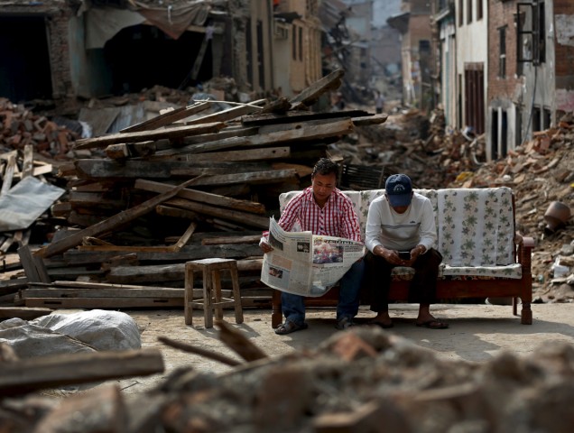 A man reads a newspaper as his friend looks at his mobile phone, as they sit next to collapsed houses on the outskirts of Kathmandu