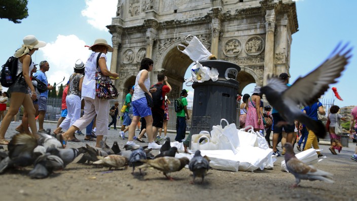 Tourists walk past the Arch of Constantine, as a pigeon flies next to a full garbage bin, in Rome