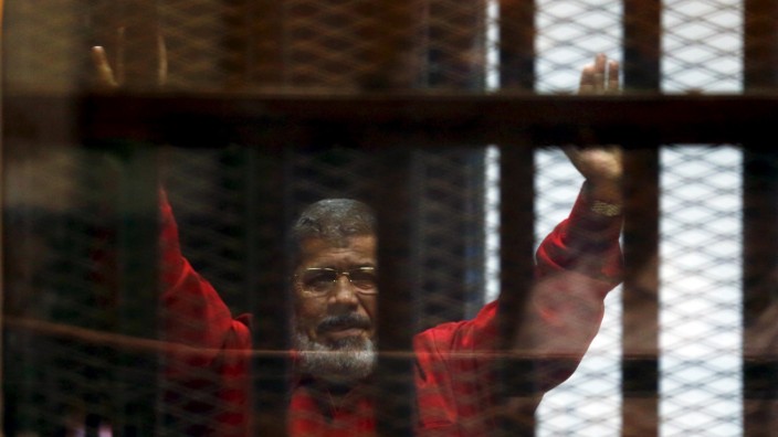Deposed President Mohamed Mursi greets his lawyers and people from behind bars at a court wearing the red uniform of a prisoner sentenced to death, during his court appearance with Muslim Brotherhood members on the outskirts of Cairo, Egypt