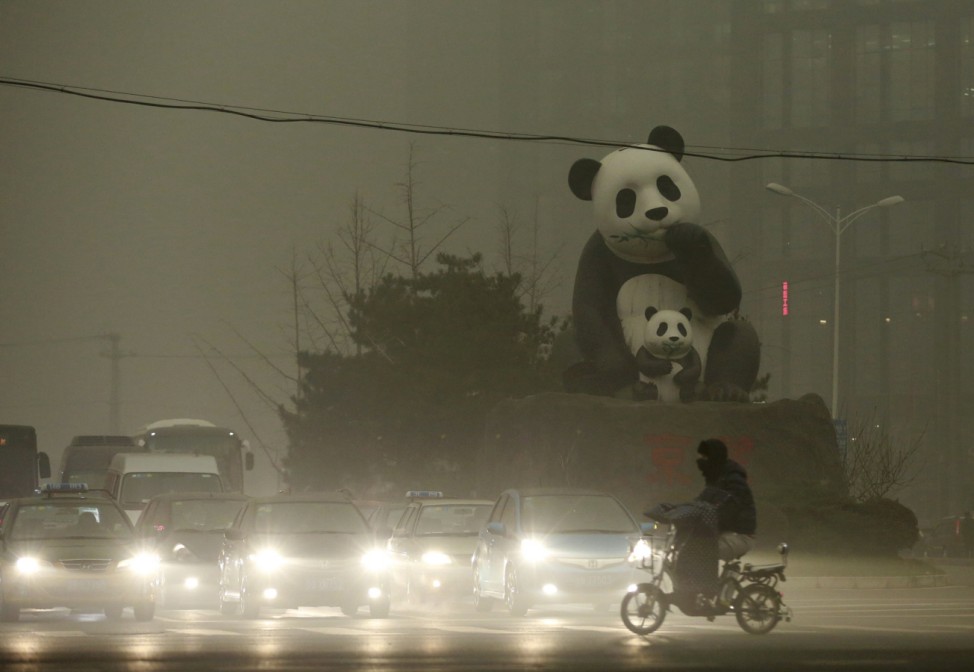 A resident rides an electric bicycle across a street amid heavy smog as vehicles wait for a traffic light next to a statue of pandas, a landmark of the Wangjing area in Beijing