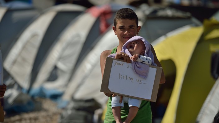 Summer School: Ein junger Flüchtling hält ein Baby und ein Schild mit der Aufschrift "Euer Schweigen tötet uns".