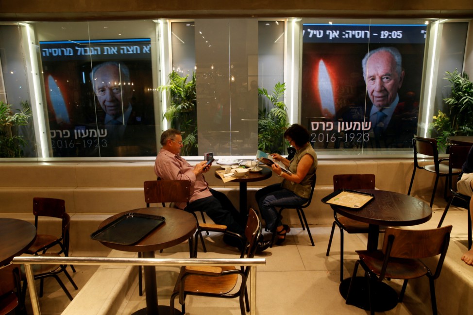People sit in a coffee shop near a wall outside a media building displaying photos of former Israeli President Shimon Peres in Tel Aviv, Israel