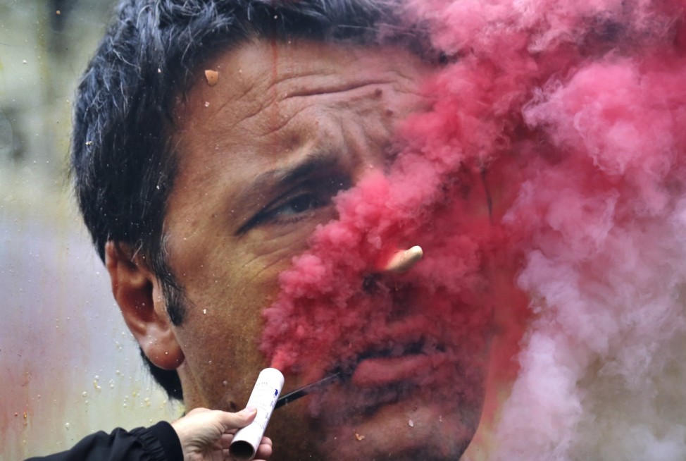 A demonstrator lights a flare in front of a board with a defaced image of Italian PM Renzi in Milan
