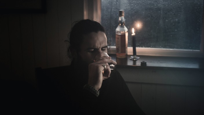 A man sitting in the dark by a window in candlelight drinkin from a small glass A bottle beside him