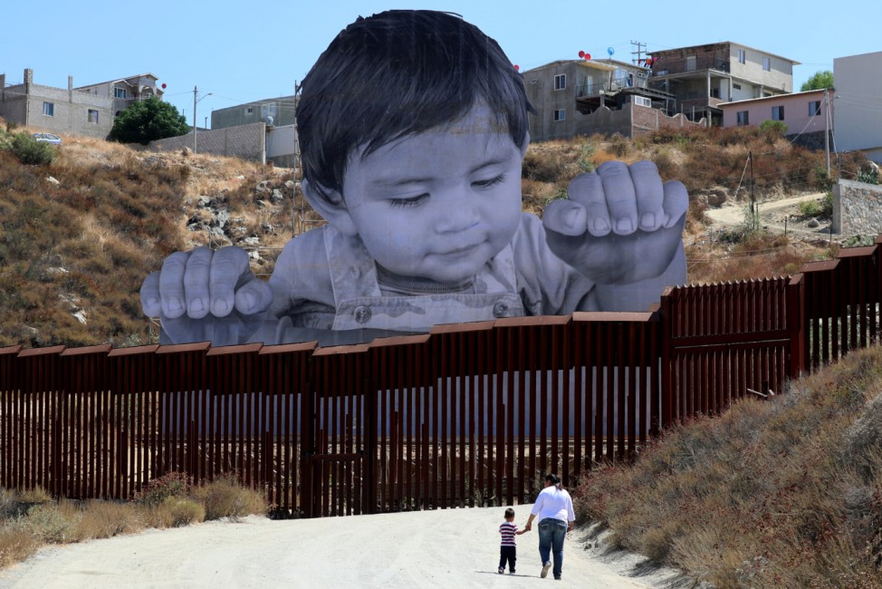 A small boy walks with his mother in front of French artist JR's image of an inquisitive baby looking into the United States over the U.S.- Mexico border wall towards Tecate, California