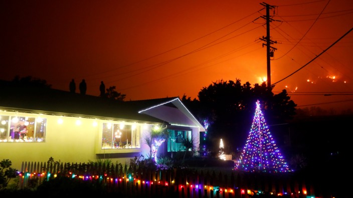 People stand on a roof of a home illuminated with Christmas lights to watch wildfire on a hillside burn during the Thomas Fire in Santa Barbara county near Carpinteria