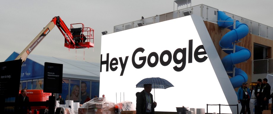 A man walks through light rain in front of the Hey Google booth under construction at the Las Vegas Convention Center in preparation for the 2018 CES in Las Vegas