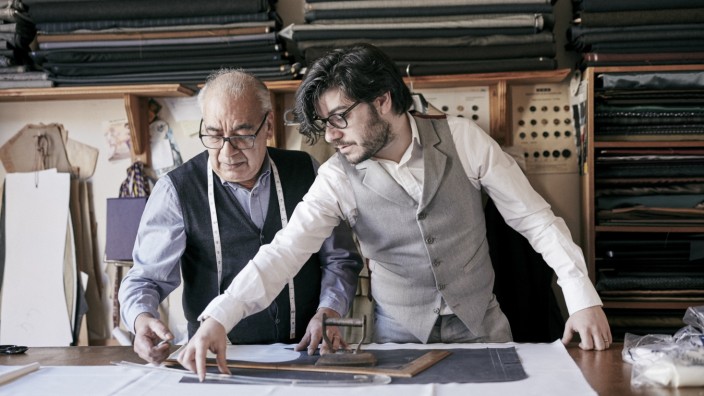 A tailor and a younger man, an assistant or apprentice working together, measuring cloth and cutting patterns.