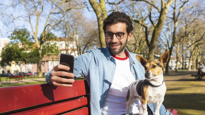 Portrait of young man sitting with his dog on park bench taking selfie with smartphone model release