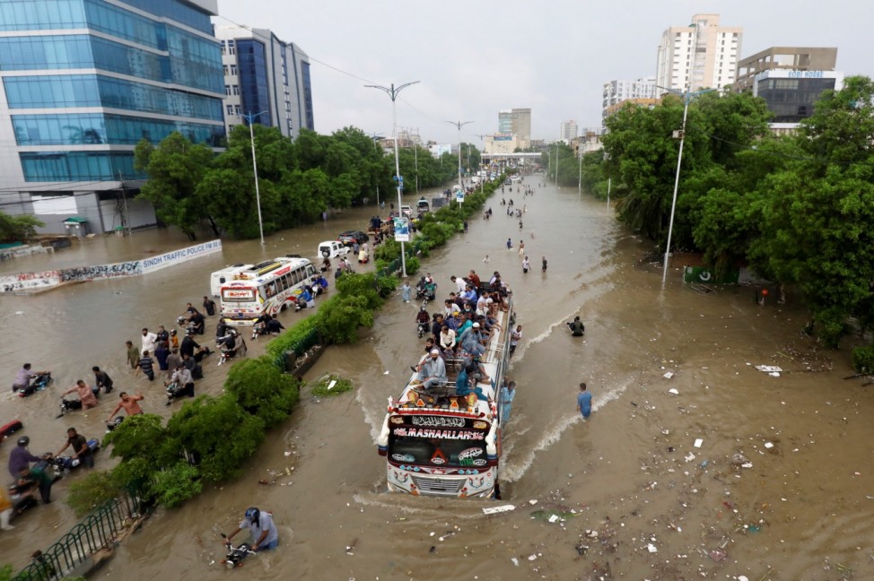 People sit atop a bus roof while others wade through the flooded road during monsoon rain, as the outbreak of the coronavirus disease (COVID-19) continues, in Karachi