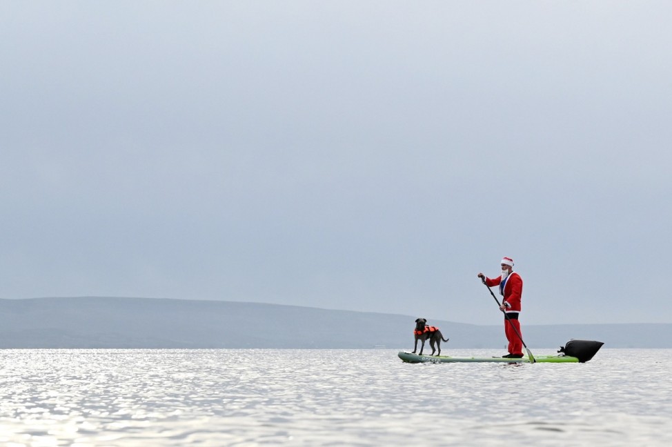 A man dressed as Santa Claus paddles on a board with his dog in Galway Bay