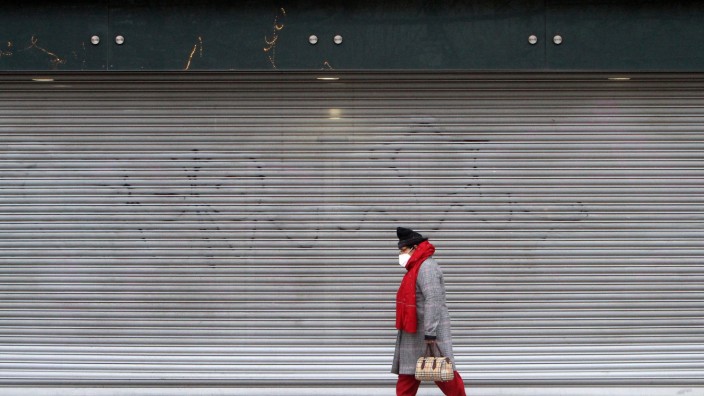 Eine Frau mit Maske geht an einer mit Rolltor verschlossenen Filiale der Modekette Primark in der Einkaufsstraße und Fu