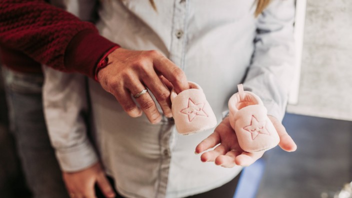 Crop young pregnant woman in casual clothes standing next to husband and together holding pink baby booties with stars,