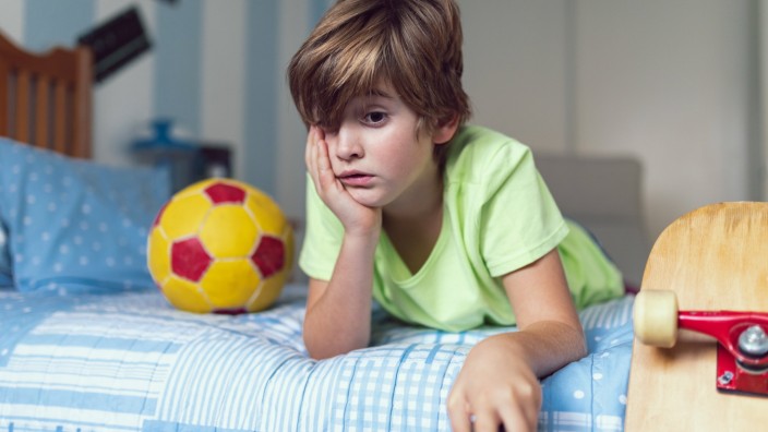 Bored little boy in casual clothes lying on bed near ball and skateboard unhappy with self isolation at home Copyright:
