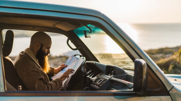 Side view of adult bearded man resting in the car and reading map at the seaside. Copyright: xx