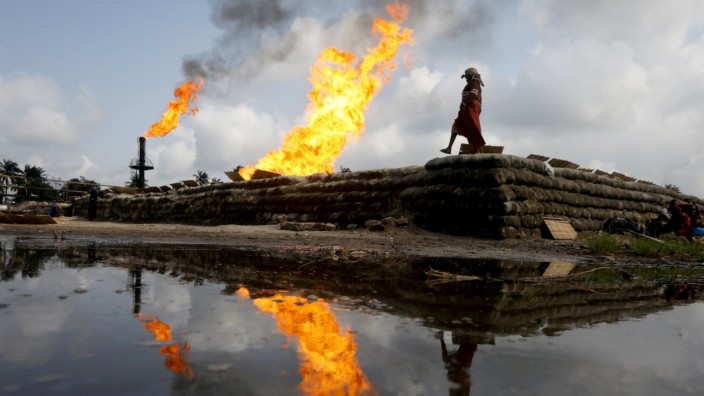 FILE PHOTO: A reflection of two gas flaring furnaces and a woman walking on sand barriers is seen in the pool of oil-smeared water at a flow station in Ughelli, Delta State