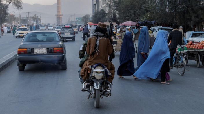 A group of women wearing burqas crosses the street as members of the Taliban drive past