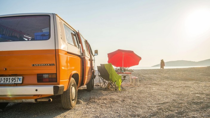 Ein Orange-weisser VW Bus Bulli steht neben einem roten Sonnenschirm am Strand des Ionischen Meer bei Saranda (Sarandë