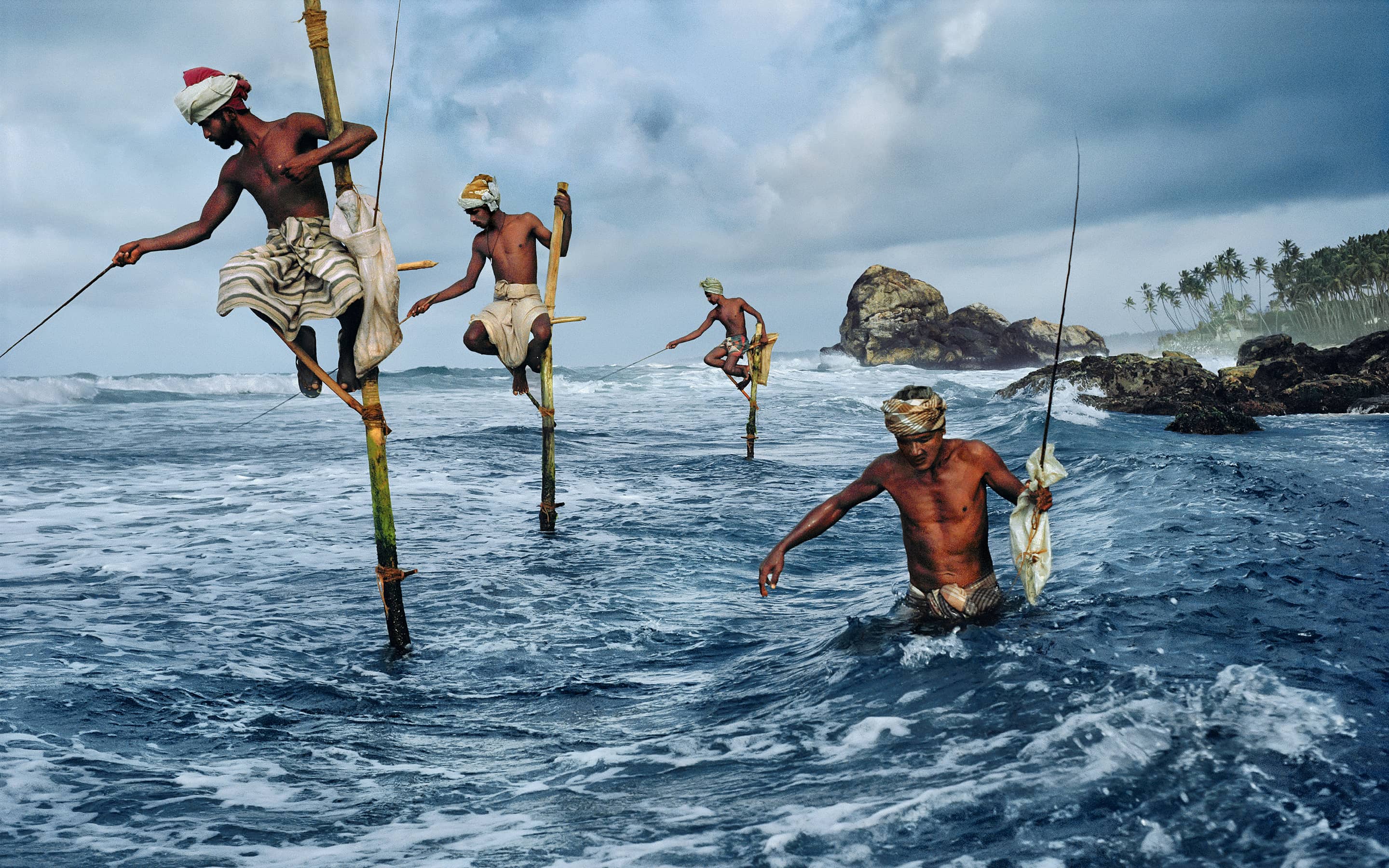 Bei Weligama an der Südküste von Sri Lanka fischen Männer im flachen Wasser auf Holzstäben kauernd, um die Beute nicht zu verscheuchen.